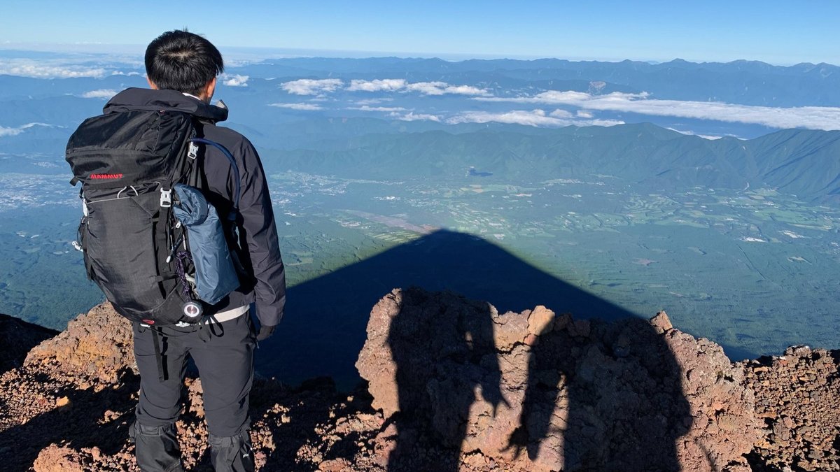 富士山頂から見下ろす雲海と影富士 レイヤリング装備で立つ登山者の後ろ姿